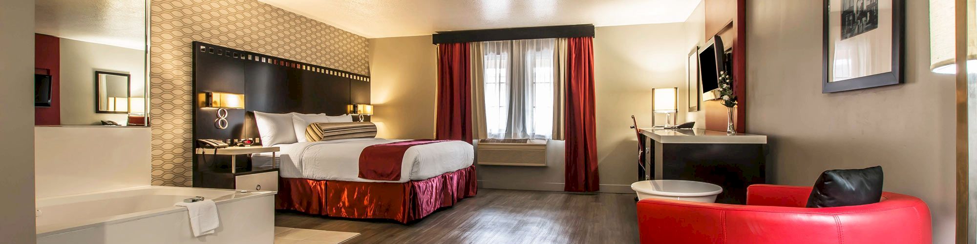 A modern hotel room with a king-sized bed, red accents, a sitting area with a red chair, a workstation, a large mirror, a bathtub, and a TV.