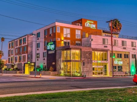 The image shows a modern, multi-story hotel building at dusk with illuminated signs reading 