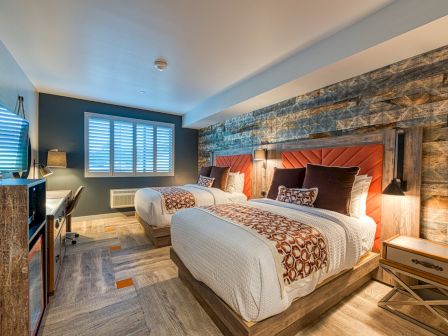 A modern bedroom with two beds, a wooden accent wall, a desk, a lamp, and a window with shutters. There’s a TV opposite the beds.