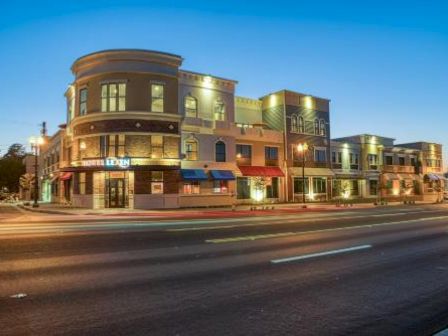 The image shows a multistory building with various storefronts illuminated in the evening, situated on a street with a few vehicles.