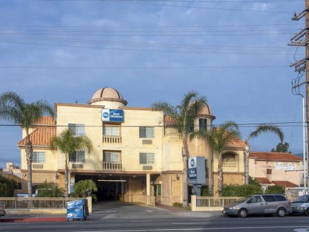 A hotel with palm trees, several parked cars, and a visible street sign. The hotel's architecture includes domes and a light yellow facade.