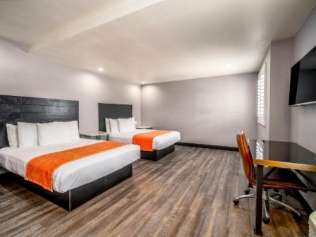 A modern hotel room with two double beds each with an orange runner, a flat-screen TV, a desk, and a chair, featuring a minimalist style and wooden floor.