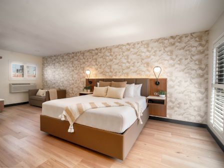 The image shows a modern, well-lit bedroom with a large bed, patterned wallpaper, bedside lamps, a sofa, wooden floors, and windows with shutters.