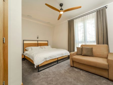 A bedroom with a bed, a beige sofa, a ceiling fan, curtains, and a window. It has a clean, minimalist design with neutral tones throughout.