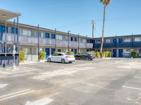 The image shows a two-story motel with a parking lot, where two cars are parked. The building has blue doors and white exteriors, with a staircase.